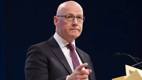 PA Media John Swinney, un hombre calvo con gafas, hace un gesto con la mano derecha mientras está de pie en el podio amarillo. Viste un traje oscuro, camisa blanca y corbata morada. 