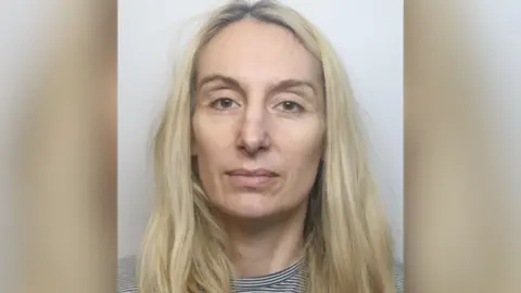 A custody shot of Gemma Kingsley. She has long blonde hair and is wearing a black and white striped top. She is looking at the camera with a blank expression.