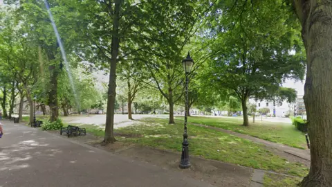 A green space with a number of mature trees and an ornate streetlight next to a pedestrian walkway