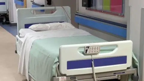 A hospital bed at Noble's Hospital, it has white plastic boards at either end and has a white pillow and sheet with pale green blanket.