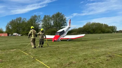 Essex County Fire & Rescue Service Two fire officers in brown and yellow uniform stand in front of a small plane which is painted white and red. The plane is facing at an angle down into the field, with its nose crumpled. There are trees behind and blue sky.