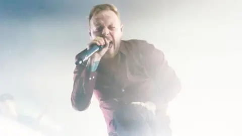 Sam Carter on stage. He's screaming into a microphone, with one leg elevated on a stage monitor in front of him. Bright white stage lights are flooding the space behind him.