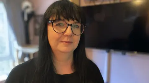 Jo Collier. She has long black hair and a fringe. She is wearing large glasses with thick brown frames. She is looking directly at the camera and smiling slightly.