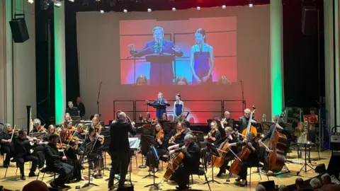 Handout An orchestra performing onstage with a man conducting them. At the back are Lorretta and Aled Jones in blue outfits. They are also projected onto a large screen above them.