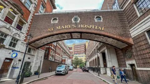Two brick buildings on either side of a road, with a bridge connecting them. The words 'St Mary's Hospital' appear on gold letters on the bridge.
