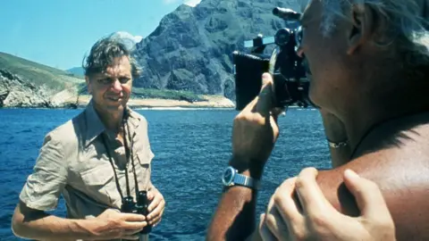 Sir David Attenborough being filmed in front of the sea and mountains for Life on Earth in 1979