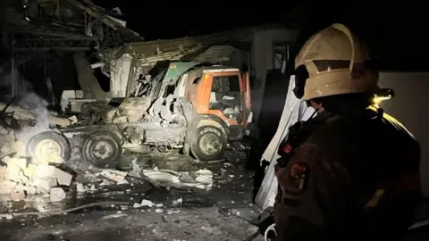 Ukrainian State Emergency Services Fire service personnel wearing a white helmet looks at a burnt out truck