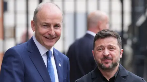 Taosieach Micheál Martin and President Volodymyr Zelensky. They are shaking hands. Martin is wearing a navy suit and Zelensky is wearing a black jacket. Martin is smiling.