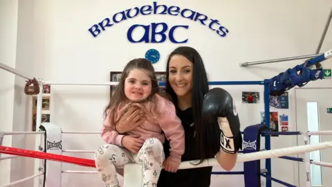 Madre con camiseta negra y con un guante de boxeo, sostiene a su hija de tres años, con polainas estampadas de tapa rosa y blanca, sentado en la cuerda superior del anillo de boxeo.