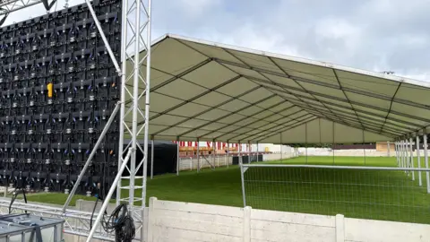 An image taken from behind a large screen with a white canopy tent stretched over a section of a football pitch.