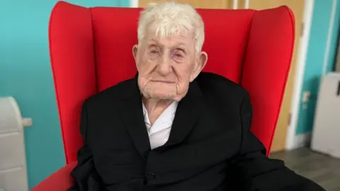 Donald Rose with short grey hair in a black blazer and white shirt sat in red armchair.
