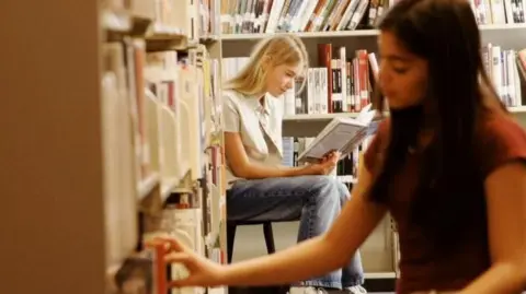 A young woman with black hair has her hand on a book on a library shelf, while a another young woman, with blonde hair and wearing a beige shirt and blue jeans, sits on a stool behind her while reading a book.