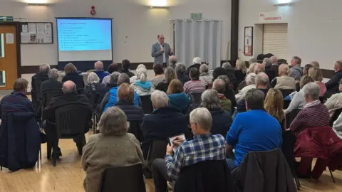 James Sherwin-Smith A public meeting with people sitting in chairs listening to a speaker who holds a microphone.