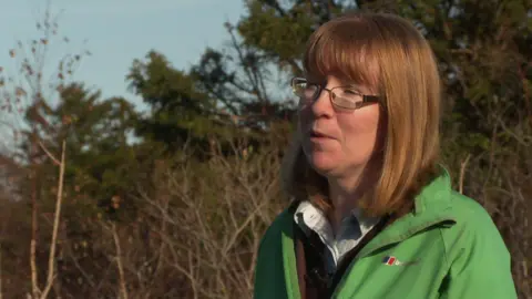 Professor Carly Stevens, wearing glasses and a green jacket, with gingery brown hair, stands in front of trees and looks off towards the left of the camera