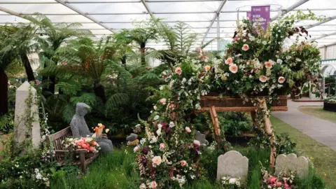 Farewell Flowers Directory A flower display with a sculpture of a man sat on a bench with a dog at his feat and an elevated willow coffin with flowers bursting out of it. Long grass and a graveyard can be seen below the coffin.