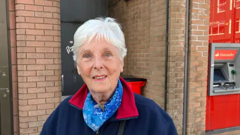 A woman with white hair is standing outside the Santander branch. Behind her to her left is a red Santander ATM. She is wearing a blue fleece and a blue scarf. She is smiling at the camera.