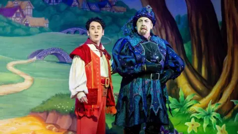 New Wimbledon Theatre A man dressed in red stands on stage beside a man dressed in blue, pulling a frown. The scene behind shows a painted background of a field with trees and winding bath.