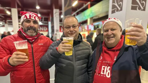 BBC Three men, two of them wearing AFC hats raising a glass a beer inside a football stadium