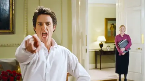 Universal Pictures / Alamy Hugh Grant, playing the prime minister, dances while pointing at the camera. He is wearing a white shirt with an open collar. Behind him a woman wearing a black skirt and a dark pink blouse, holding a file under her arm, watches him.