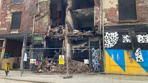 A brick building with the front collapsed into the street it is surrounded by mental fences and 'danger' signs.