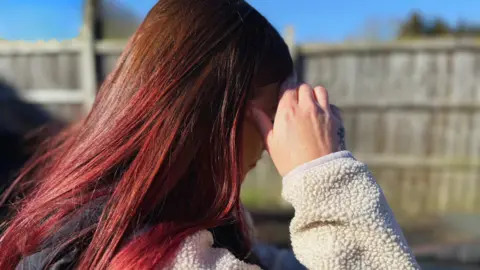 A side profile of Corrina with dyed red hair, looking at the ground and shielding her face with her right hand. 