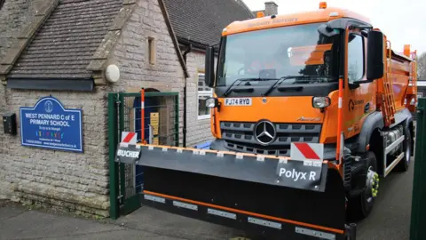 An orange gritter rolling out of a school gate. The school building next to the gate is made of grey stone and has a plaque on the wall with white writing on a blue background