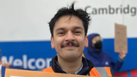 Alex Dunlop/BBC Dr Nick Singh is in the middle. He has short black hair and a moustache, and is wearing an orange hi-vis and is stood in front of a welcome board. 