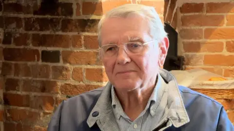 Richard Brown gives a TV interview indoors with an exposed brick wall behind him. He has white hair and glasses and wears a casual jacket over a striped shirt.