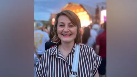 Alex Pope/BBC Carole Soraghan with the crowd behind her at dusk. She wears a navy and white striped blouse. She is smiling and a bag strap is across her chest. 