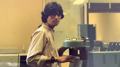 Cancer Research UK Old photograph of Peter with dark hair and glasses wearing and shirt and standing in a laboratory with his hands touching some equipment