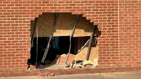 A large hole can be seen in a brick wall where a car smashed through it. Bricks and rubble can be seen inside the hole.
