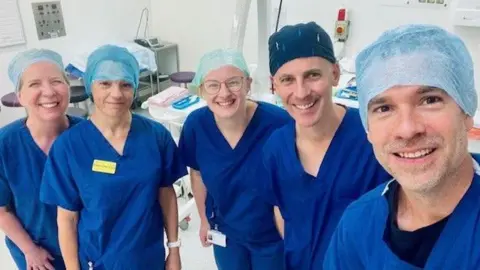 KING'S COLLEGE HOSPITAL NHS FOUNDATION TRUST The team of medical professionals involved on the day. They are wearing blue scrubs and are standing in a clinical area of a hospital smiling to camera