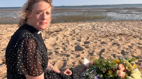 A woman with blonde curly and wavy hair kneels on a sandy beach looking at the camera. She wears black including a white dog collar. Behind her is the sea. Beside her is a bouquet of flowers.