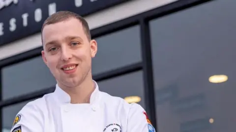 Weston Grove handout Close up of James Ellams with short brown hair wearing chef whites is standing outside the Weston Grove fish and chip shop. He is folding his arm. He is smiling.