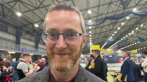 Philip McGuigan has short brown hair which is spikey at the front and a short ginger beard. He is wearing glasses and a tweed grey jacket and is standing in an election count centre.