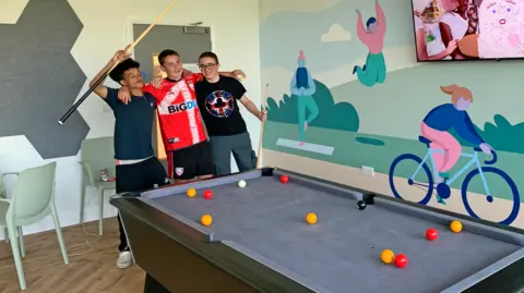 Three teenage boys, smiling and arm-in-arm beside a pool table in a room with a stylised image painted on the wall in the background of people enjoying yoga, cycling and jumping