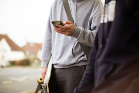 A teenager holding a skateboard uses his smartphone