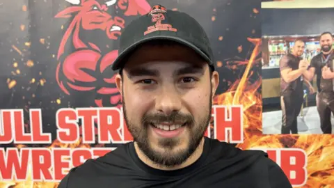 A man with a black t-shirt, a black cap, and a beard