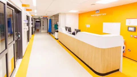 PHU Adult Urgent Care at Queen Alexandra Hospital, showing a white and bright yellow corridor with a reception desk on one side.