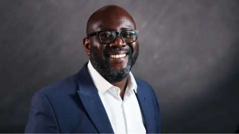 Chewe Munkonge headshot. He is bald, with dark facial hair and glasses. He is wearing a blue blazer over a white shirt and is standing in front of a grey wall.