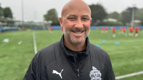 Phil Parkinson is bald with a grey beard, he smiles and wears a dark rain coat. He stands by a green football pitch with players wearing red blurred out behind him.