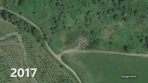 Google Earth A satellite image with 2017 in white blocks on the bottom left of the image. The image shows fields with trees a single track road running through the countryside with no clear issues with this countryside scene. There seems to be some white dots at the entrance to the site which has allegedly been used for waste dumping but it's difficult to make out what they are.