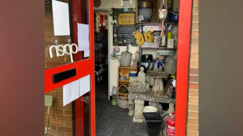 Laura Foster/BBC A red glass door is open and inside is lots of stock which includes shelves containing signs, garden ornaments, a metal watering can.