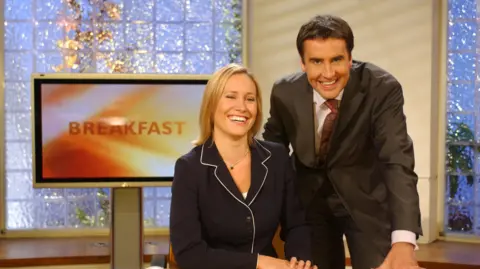 Dermot Murnaghan and Sophie Raworth smile at the camera while stood in front of a TV that says Breakfast in 2002.