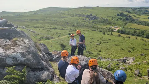 Four girls in helmets watch their friend abseil down a rock face with the help of an outdoor instructor.