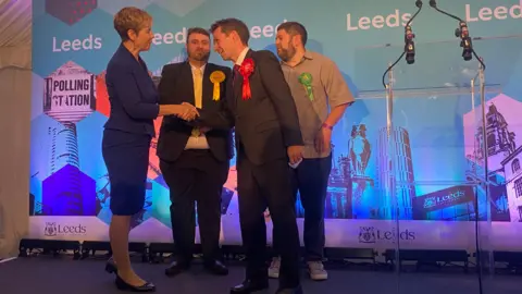 Mark Sewards shakes hands with Andrea Jenkyns at an election count. Behind them are candidates for the Liberal Democrats and the Green Party.