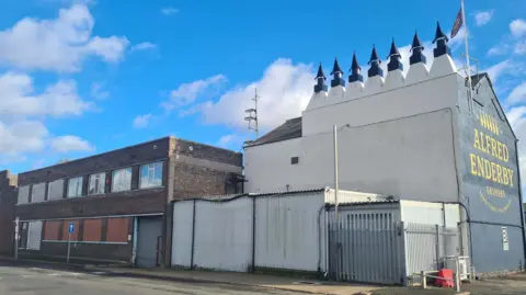 The Alfred Enderby smokehouse building topped by a row of seven ornate chimneys. Part of the building is white and the side is blue, with the words Alfred Enderby written on it in yellow.