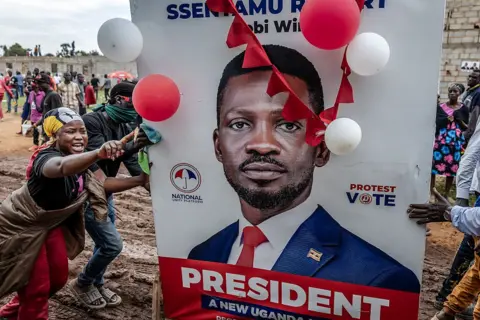 AFP via Getty Images A large poster of Bobi Wine draped in red and white balloons dominates the picture. On the left of the image a woman is shouting and waving her finger.