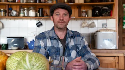 Will is sitting at a kitchen table and wearing a dark flat cap and blue and white chequered shirt. He has short brown hair. Behind him is a wooden shelving unit with various jars of ingredients, mugs, a toaster, a teapot and a breadbin upon it. In front of him is a glass of water and a fruit bowl with fruit inside.