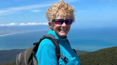 Liz Odell smiling at the camera while hiking up a mountain. She has short blonde curly hair and is wearing purple polarised mountaineering sunglasses, a blue half-zip fleece and a black back park.
